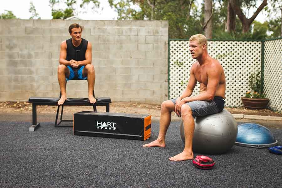Two men training outdoors: one sits on a bench while the other rests on a large exercise ball, with workout equipment and a Hart Sports plyobox nearby.