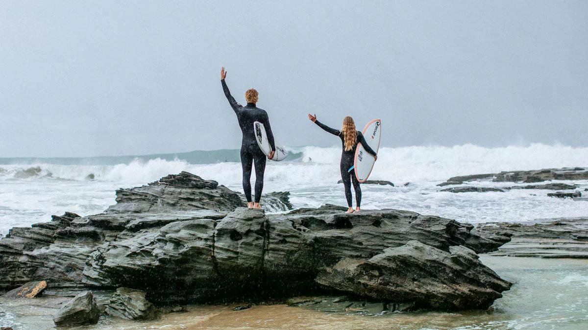 Two surfers stand on a rocky outcrop near the sea, holding their boards and waving to the horizon.