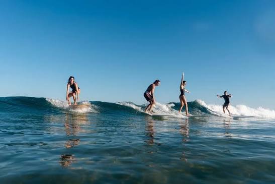 Vier Surferinnen reiten kleine Wellen, klarer Himmel, Wasserreflexe unten.