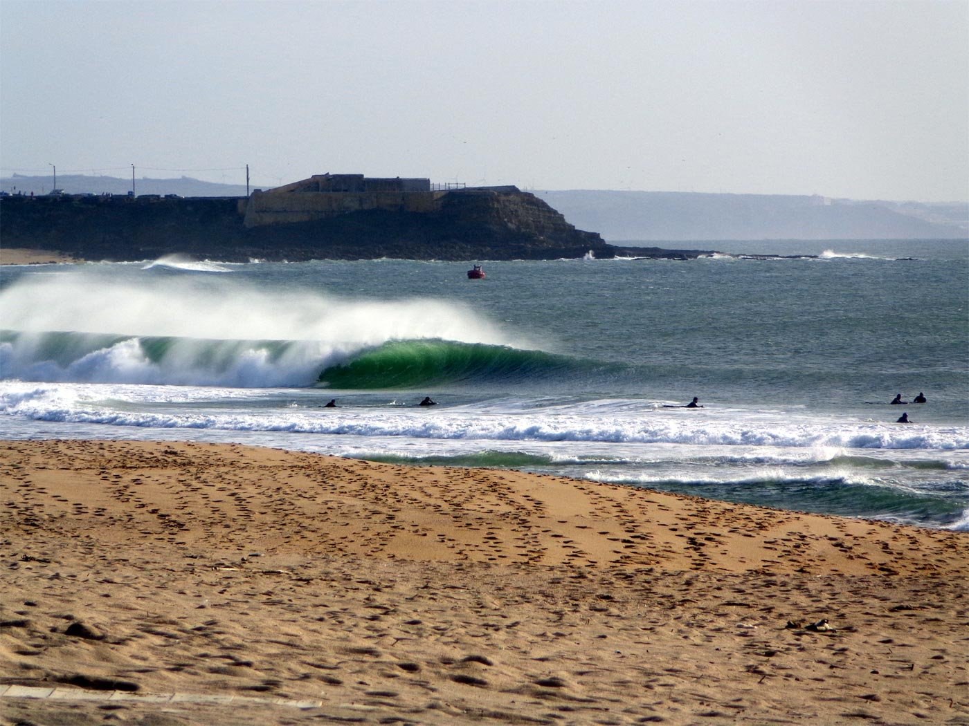 Wellen brechen nah am Sandstrand, einige Surfer im Wasser, Küste mit Felsen im Hintergrund.