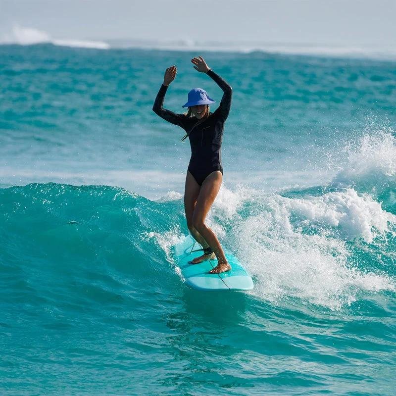 Woman surfing turquoise waves on a light-blue board, wearing a black wetsuit and blue hat, balancing with arms raised.