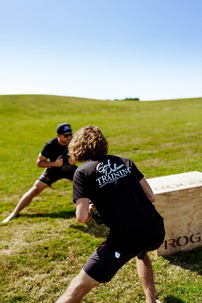 Zwei Sportler beim Outdoor-Training auf grünem Hügel, einer wirft einen Haltblock.