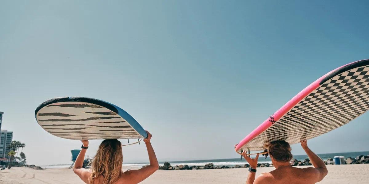 Zwei Surfbretter werden am Strand quer über die Köpfe getragen, im Hintergrund Meer, Strand und blauer Himmel.